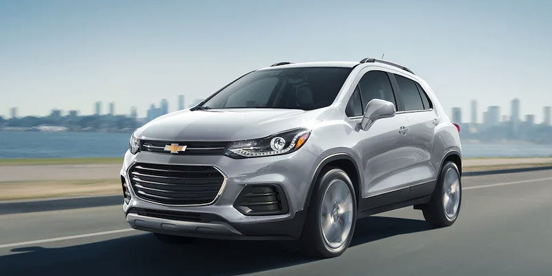 Silver Chevrolet Trax driving on a road near a waterfront skyline.